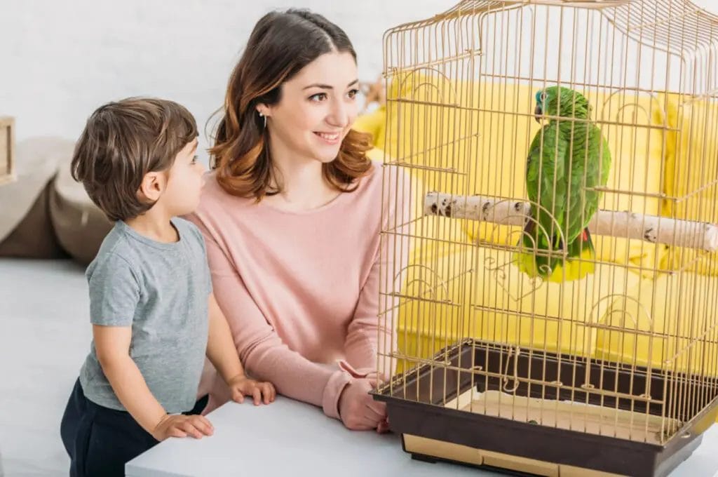 A woman and a young child look at a green parrot inside a cage placed on a table, contemplating the parrot purchase.